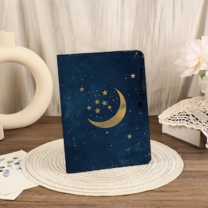 Può includere: Un diario blu scuro con un design a luna crescente e stelle dorate. La copertina ha un aspetto strutturato con un tema celeste. Il diario è esposto su un sottobicchiere intrecciato.