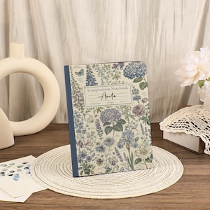 May include: A composition notebook with a cream-colored cover featuring a botanical print of blue and purple flowers, butterflies, and green foliage. The center label reads Composition Notebook, Amelia, College Ruled, 100 Pages, 8.5 x 11.