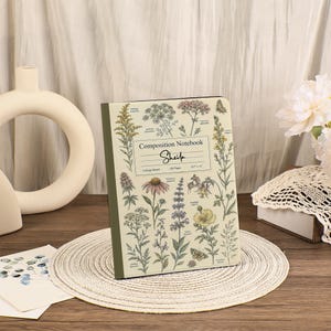 May include: A composition notebook with a cream-colored cover featuring various botanical illustrations of wildflowers and butterflies. The center label reads Composition Notebook, Sheila, College Ruled, 120 Pages, 8.5 x 11 inches.