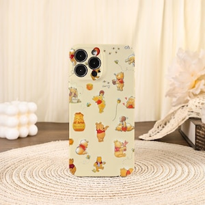 May include: A cream-colored phone case featuring a repeating pattern of Winnie the Pooh characters in various poses. The case has a matte finish and includes the camera lens cutouts. Honey pots and bees are also part of the design.