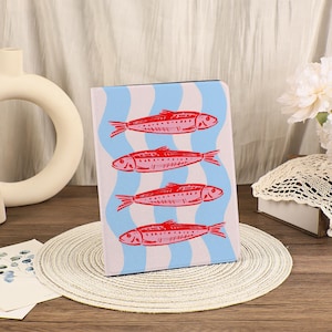 Könnte beinhalten: Ein Notizbuch mit einem sich wiederholenden Muster aus roten Fischen auf einem hellblauen und weißen wellenförmigen Hintergrund. Die Fische sind stilisiert mit detaillierten Flossen und Schuppen. Das Notizbuch wird auf einem runden, gewebten Platzset präsentiert.