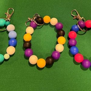 May include: Three beaded keychains in various color combinations. One features blue, green, and cream beads. Another has purple, black, and yellow beads. The third keychain has red, purple, and blue beads. All have gold-colored clasps.