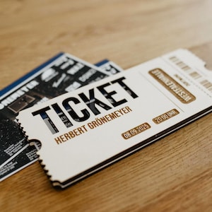 Könnte beinhalten: Ein weißes Holzticket mit dem schwarzen Schriftzug TICKET und HERBERT GRÖNEMEYER. Das Ticket enthält goldfarbene rechteckige Akzente mit dem Text WESTFALENHALLE, 08.09.2025 und 20:00 UHR. Ein Strichcode ist in der oberen rechten Ecke zu sehen.