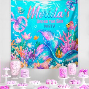 The image is a vibrant underwater-themed party backdrop with a mermaid tail and various marine life, including fish, coral, and sea anemones. The backdrop is set against a blue ocean backdrop, and the text "Merida's Under the Sea Party" is prominently displayed. In front of the backdrop, there is a table filled with an assortment of colorful candies and sweets, creating a festive atmosphere for the mermaid-themed party.