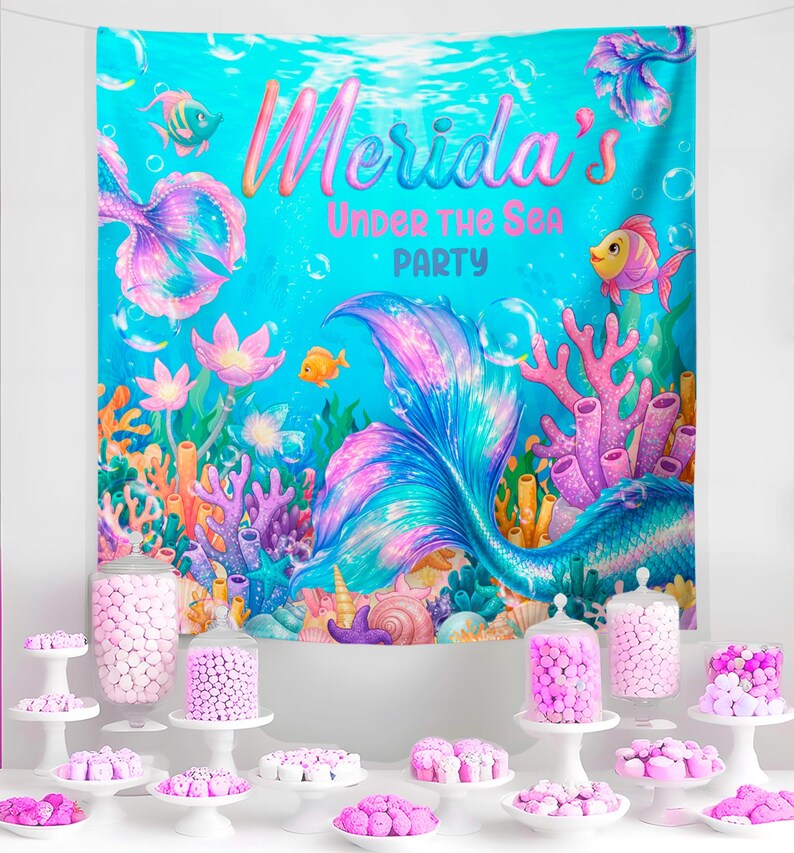 The image is a vibrant underwater-themed party backdrop with a mermaid tail and various marine life, including fish, coral, and sea anemones. The backdrop is set against a blue ocean backdrop, and the text "Merida's Under the Sea Party" is prominently displayed. In front of the backdrop, there is a table filled with an assortment of colorful candies and sweets, creating a festive atmosphere for the mermaid-themed party.