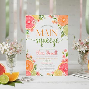 May include: A bridal shower invitation with a citrus theme. The invitation features the text "She found her Main Squeeze" in orange and green, with the name "Olivia Bennett" in pink. The design includes illustrations of citrus fruits and flowers.