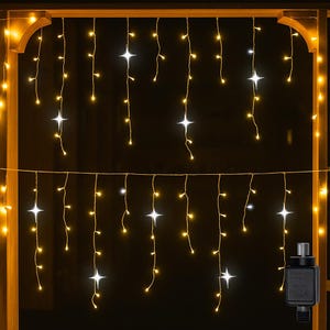 Op de afbeelding: Slingers met warmwitte en stervormige lampjes die aan een houten frame hangen. De lampjes zijn gerangschikt in verticale strengen, waardoor een gordijneffect ontstaat. Een zwarte voedingsadapter is zichtbaar aan de rechterkant.