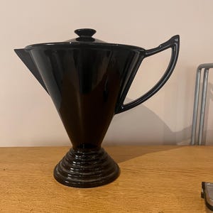 May include: A glossy black ceramic teapot with a wide, flared top, a pointed spout, and a triangular handle. The teapot features a matching lid and sits on a circular, tiered pedestal base. The design has a smooth, reflective finish throughout.