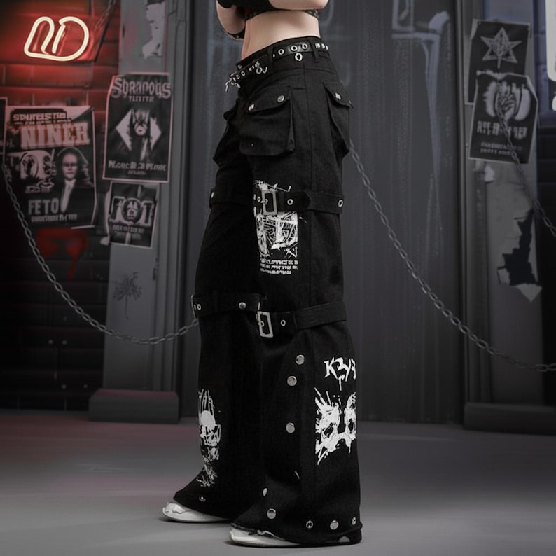 May include: Black cargo pants with white graphic designs, buckles, and rivets. The pants have multiple pockets and a wide leg design. The overall style is edgy and alternative.
