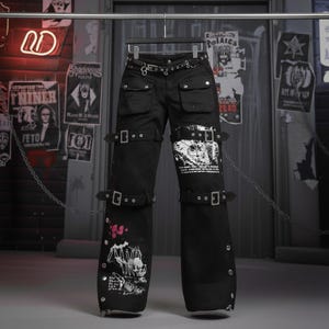 May include: Black pants with multiple pockets, straps, and buckles. The pants have a wide leg design and feature graphic prints in white and pink. A black belt with silver hardware is included.
