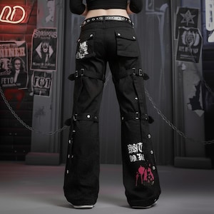 May include: Black cargo pants with multiple straps, buckles, and pockets. The pants have white text and a pink graphic on the lower leg. The pants are worn with a black belt and are part of a punk or alternative fashion style.