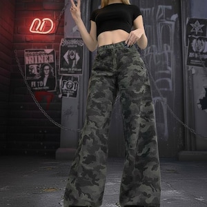 May include: A person wearing a black crop top and camouflage print wide-leg jeans. The jeans have a green, gray, and black pattern. The person is wearing black combat boots. The background includes graffiti and posters.