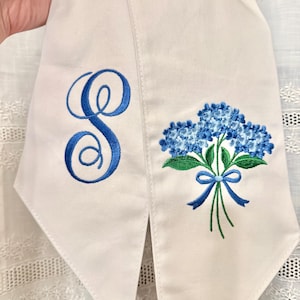 May include: White graduation stole with blue embroidered details. One side features a large, cursive letter "S". The other side displays a bouquet of blue hydrangeas with green leaves and a blue ribbon.