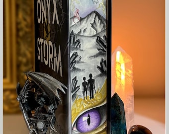 Könnte beinhalten: Ein Buch mit dem Titel "Onyx Storm" mit einem schwarzen Einband und einem Drachen-Design auf dem Rücken. Der Rücken zeigt einen Drachen mit violetten Augen, Blitzen und einer Bergszene. Das Buch ist gegen eine silberne Drachenfigur gelehnt.