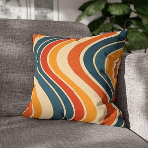 Puede incluir: Cojín cuadrado con un patrón retro ondulado en tonos naranja, rojo, amarillo y azul verdoso. El cojín tiene un fondo crema y está colocado sobre un sofá gris. El diseño del cojín evoca una estética vintage de los años 70.