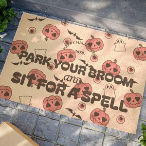 Op de afbeelding: Beige Halloween deurmat met een herhalend patroon van pompoenen, vleermuizen, ogen en geesten. De tekst "PARK YOUR BROOM and SIT FOR A SPELL" is gedrukt in donkerbruin. De woorden "HAPPY HALLOWEEN 1978" staan ook op de mat.