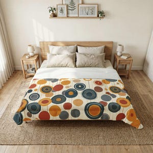May include: A bed with a wooden headboard and two bedside tables. The bed is covered with a cream-coloured blanket featuring a pattern of colourful circles in shades of orange, blue, grey, and red. The room has a neutral colour palette with framed artwork on the wall.