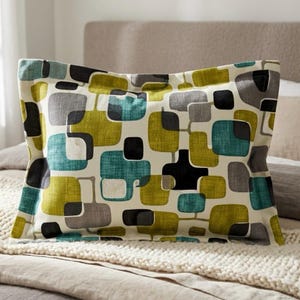 May include: Rectangular pillow with a cream background and a repeating pattern of geometric shapes in teal, olive green, gray, and black. The pillow is placed on a bed with a neutral-colored blanket and a beige headboard.