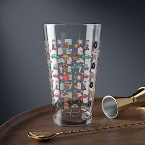 Puede incluir: Una coctelera de vidrio transparente con un diseño retro de temática de restaurante. El diseño incluye imágenes de coches clásicos, máquinas de discos, discos y personas. Una cuchara y un jigger dorados están en una bandeja de madera.