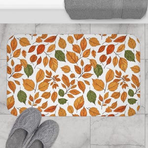 May include: Rectangular bath mat with a white backdrop, adorned with a pattern of autumnal leaves in orange, yellow, and green hues. The leaves are spread across the mat, creating a seasonal design. Grey slippers are at the bottom.