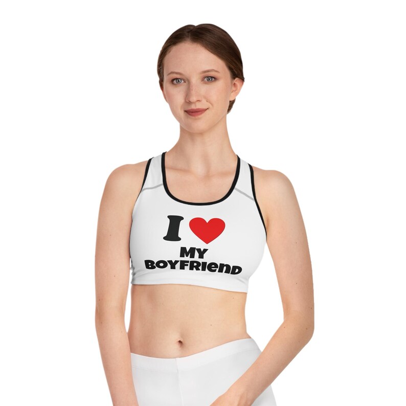 May include: White sports bra with black trim and the text "I &hearts; MY BOYFRIEND" in black and red. The bra has a racerback design and is made of a soft, stretchy material. The bra is designed for athletic activities.
