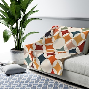 May include: A colorful throw blanket with a geometric pattern of diamonds and circles in shades of orange, red, blue, and cream. The blanket is draped over a gray sofa, creating a cozy and stylish look. A large potted plant is in the background.