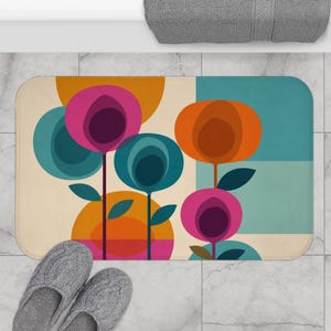 Puede incluir: Una alfombrilla de baño rectangular con un patrón floral gráfico de flores circulares estilizadas en tonos magenta, morado oscuro, azul cerceta y naranja. El diseño incluye bloques de color sólido y tallos finos sobre un fondo crema.