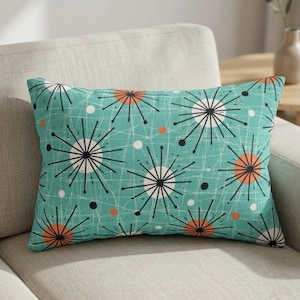 Op de afbeelding: Rechthoekig decoratief kussen met een turquoise achtergrond en een mid-century modern sterrenpatroon in zwart, wit en oranje. Het kussen ligt op een lichtgekleurde bank. Het ontwerp bevat witte lijnen en kleine witte en oranje stippen.