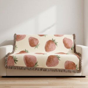 Könnte beinhalten: Eine cremefarbene Decke mit einem sich wiederholenden Muster aus großen, rosa Erdbeeren mit grünen Blättern. Die Decke hat einen Fransenrand und ist über ein weißes Sofa drapiert, was der Raumdekoration einen Hauch von Laune verleiht.