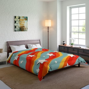 Può includere: Una trapunta colorata con un design retrò. Presenta strisce ondulate rosse, arancioni, gialle e blu, con accenti di stelle bianche. La biancheria da letto è su una struttura letto in legno in una stanza ben illuminata.
