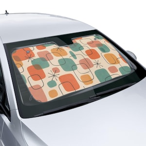 May include: A car windshield sunshade with a retro design. The pattern features orange, teal, and pink squares and starbursts on a cream background, outlined in black. The sunshade is installed in a silver car.