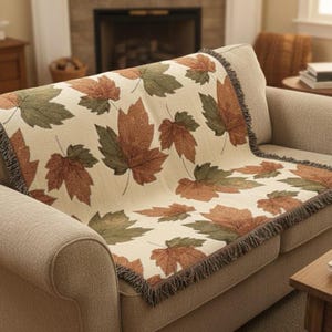 May include: A beige throw blanket with a repeating pattern of large maple leaves in shades of brown and green. The blanket is draped over a beige sofa and has a fringed edge.
