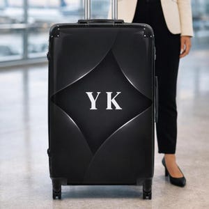 Könnte beinhalten: Schwarzer Rollkoffer mit den Initialen "YK" in Weiß. Der Koffer hat ein geometrisches Design auf der Vorderseite und einen ausziehbaren Griff. Der Koffer steht auf einer glatten Oberfläche.