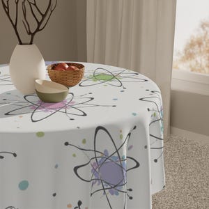 May include: A white round tablecloth with a colourful atomic design. The design features black lines and pastel colours, including pink, green, and purple. A white vase, a small woven basket with fruit, and two bowls are on the table.