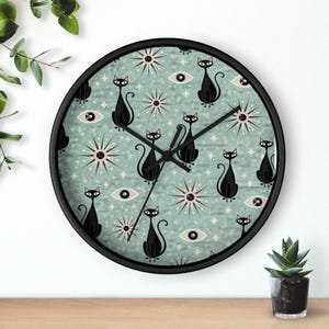 May include: A round wall clock with a black frame. The clock face features a repeating pattern of black cat silhouettes, stylized eyes, and starbursts on a teal background. The clock hands are black.
