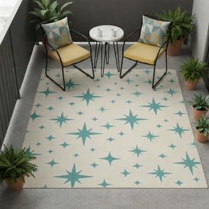 May include: An outdoor patio scene featuring a cream-colored rug with a teal starburst pattern. Two yellow-cushioned chairs with black metal frames and a small white table are set on a concrete patio. Potted plants add greenery to the space.