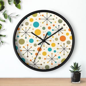 Pode incluir: Um relógio de parede redondo com moldura preta. O mostrador do relógio apresenta um fundo creme com um padrão de círculos coloridos e desenhos de estrelas em laranja, amarelo, azul e verde. Os ponteiros pretos indicam as horas.