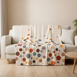 May include: A cream-coloured throw blanket draped over a sofa. The blanket features a pattern of vertical brown stripes and colourful circles in shades of brown, orange, blue, and yellow. The sofa has a neutral colour and a matching pillow.