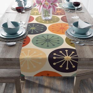 May include: A table runner with a retro design featuring large, colorful circles in shades of orange, green, and purple on a cream background. The runner is placed on a wooden table set for a meal, with plates, glasses, and flowers.