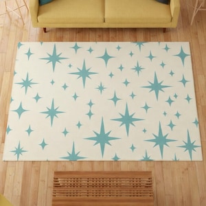 May include: A rectangular rug featuring a cream-coloured background adorned with a repeating pattern of teal starbursts. The rug is placed on a wooden floor, with a yellow sofa visible in the background.