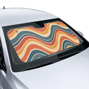 Op de afbeelding: Een auto voorruit zonnescherm met een retro golvend patroon in tinten oranje, blauw en crème. Het zonnescherm is geïnstalleerd in een zilveren auto en biedt bescherming tegen de zon.