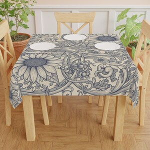 May include: A rectangular tablecloth with a detailed floral design, including sunflowers and other botanical elements in blue and cream. The tablecloth is on a wooden table, with white plates. The table is 120 cm x 120 cm.