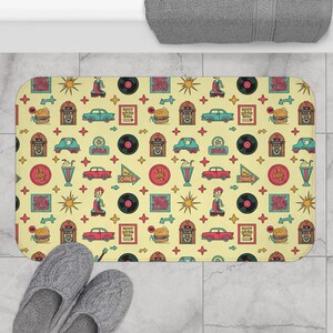 May include: A rectangular bath mat with a retro diner theme. The mat features a repeating pattern of classic cars, jukeboxes, burgers, vinyl records, and diner signs on a yellow background. Gray slippers are in the foreground.