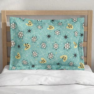 May include: A turquoise pillowcase with a retro atomic pattern. The design features white lines, black stars, and yellow and white atomic shapes. The pillowcase is displayed on a wooden bed frame.