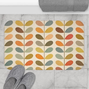 Può includere: Un tappetino da bagno rettangolare con sfondo crema e un motivo ripetuto di forme colorate a forma di foglia in tonalità arancione, giallo, marrone e verde. Due pantofole grigie a maglia sono in basso.