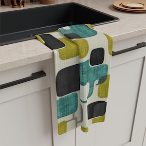 May include: A kitchen towel with a geometric pattern in teal, black, and chartreuse green on an off-white background. The towel is hanging over the edge of a black sink, with white cabinets and black hardware visible.