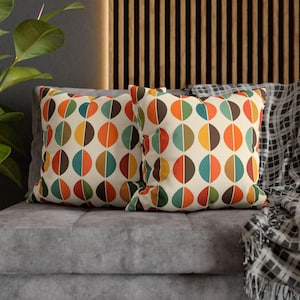 May include: Two square cushions featuring a cream backdrop and a repeating pattern of colourful half-circles in orange, brown, green, and yellow. The cushions are on a grey sofa, with a patterned blanket visible.