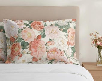 Puede incluir: Fundas de almohada con un diseño floral de rosas rosas, melocotón y blancas con follaje verde sobre un fondo crema. Las fundas de almohada están sobre una cama blanca, con un cabecero beige.