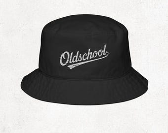 May include: Black bucket hat with the word "Oldschool" in white, stylized script. The hat has a wide brim and a classic, casual design. The text is slightly distressed, giving it a vintage look.