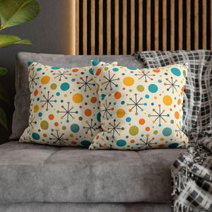 Peut inclure: Deux coussins carrés avec un fond crème, ornés d'un motif coloré de cercles orange, jaunes, turquoise et verts, et de motifs en forme d'éclats noirs. Les coussins sont posés sur un canapé gris.